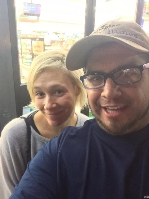 Two people smiling at the camera indoors near a window